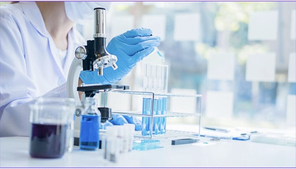 Picture of a woman in a lab coat with a face mask and gloves using a pipette in a lab setting with blue liquids in small flasks. 